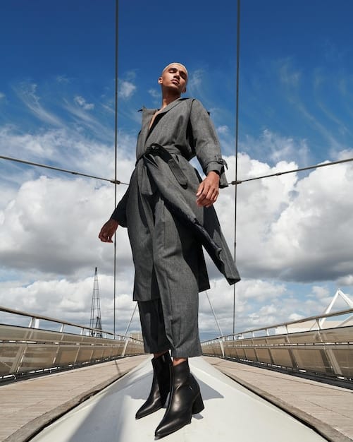 A full-length shot of a model wearing a modern interpretation of a classic trench coat. The trench coat is deconstructed with exaggerated sleeves and a unique asymmetrical cut. The model is wearing it over a simple black dress and ankle boots.
