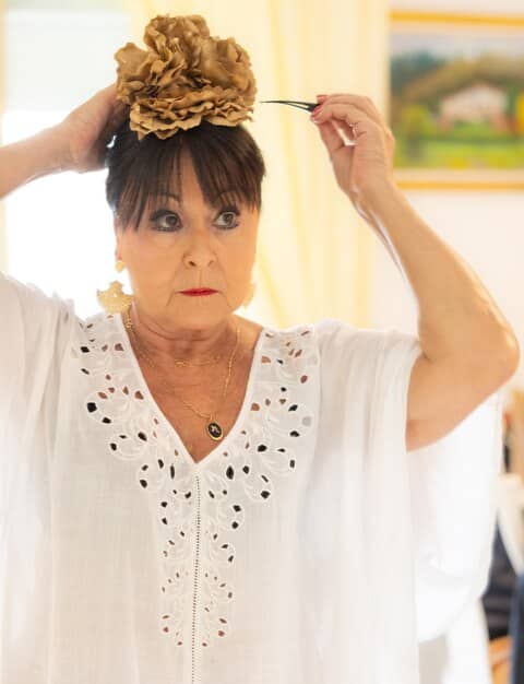 A close-up of a woman in France quickly styling her hair into a sleek updo. She's wearing a simple blouse, but her hair transformation and statement earrings hint at an evening event. The background is a modern apartment with natural light.