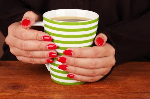A woman's hand holding a cup of coffee, showcasing a perfectly manicured set of nails painted with a shimmering coral nail polish. The background features a vibrant outdoor cafe setting, with colorful flowers and sunlit tables. The image highlights the cheerful and summery vibe of the coral nail polish.