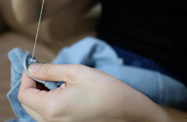 A close-up shot of a hand reinforcing the stitches of a button on a piece of fabric using a needle and thread. The focus is on the stitching technique and the details of the fabric.
