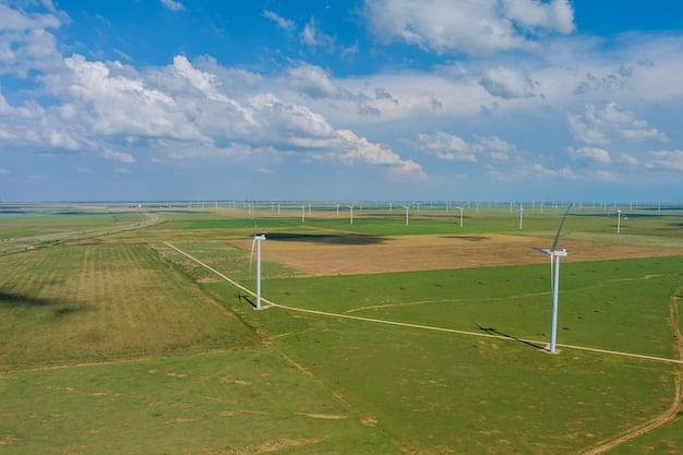 An aerial view of a large solar panel farm in a rural area, with wind turbines visible in the distance, set against a clear blue sky.
