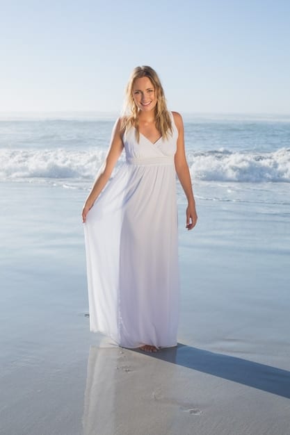 A woman with an hourglass figure standing on a sunny beach, wearing a flowy maxi dress with a floral print. She is accessorized with a wide-brimmed sun hat and sandals. The overall feel is relaxed and stylish.