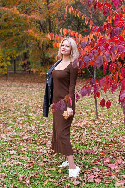 A woman with a pear-shaped figure walking through a park with colorful fall foliage, wearing a fitted turtleneck sweater, a midi skirt, tights, and ankle boots. She is carrying a leather tote bag. The look is sophisticated and reflects the autumnal colors.