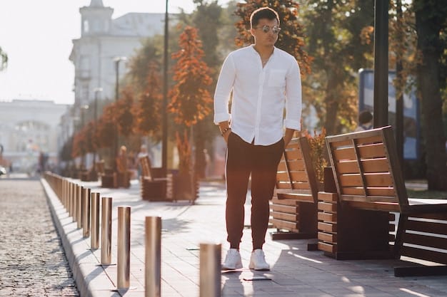 A man wearing a light blue linen shirt, khaki chinos, and white sneakers, walking down a sunny street with blooming trees in the background.