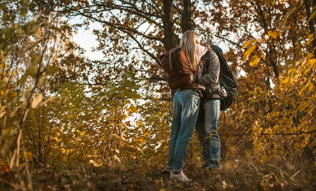 A man in a brown leather jacket, dark jeans, and boots, walking through a park with trees showing fall foliage.